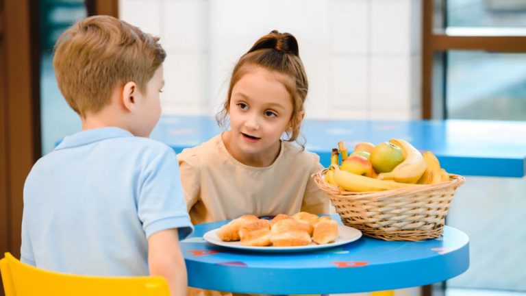 Two children engaging in friendly conversation over breakfast with fruit and pastries.