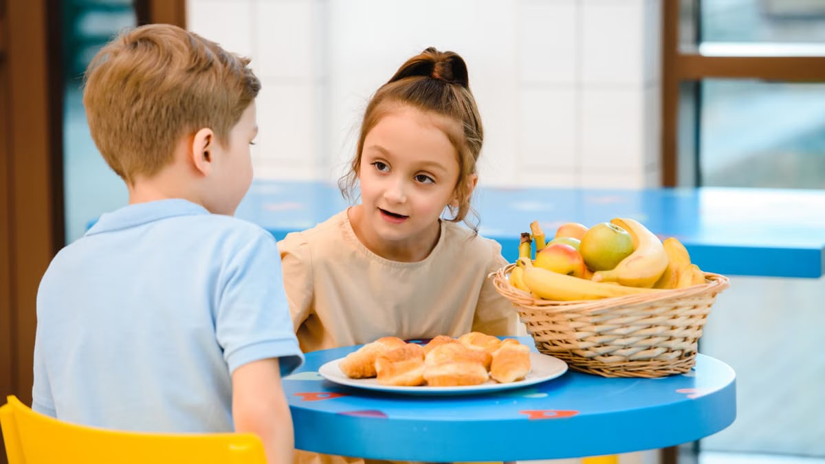 Two children engaging in friendly conversation over breakfast with fruit and pastries.